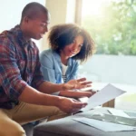 Casal lendo alguns papéis com laptop aberto na sala de casa, representando o empréstimo com garantia de imóvel para negativado.
