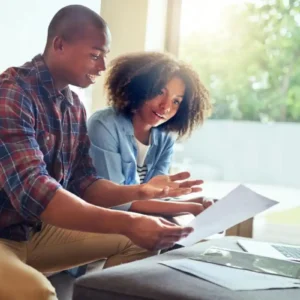 Casal lendo alguns papéis com laptop aberto na sala de casa, representando o empréstimo com garantia de imóvel para negativado.