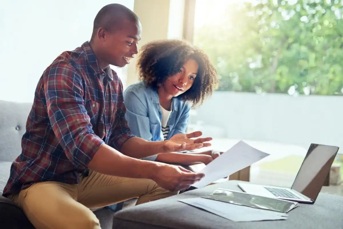 Casal lendo alguns papéis com laptop aberto na sala de casa, representando o empréstimo com garantia de imóvel para negativado.
