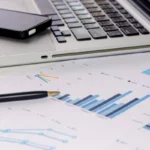 Celular, laptop, papéis e caneta sobre uma mesa, representando produtos financeiros.