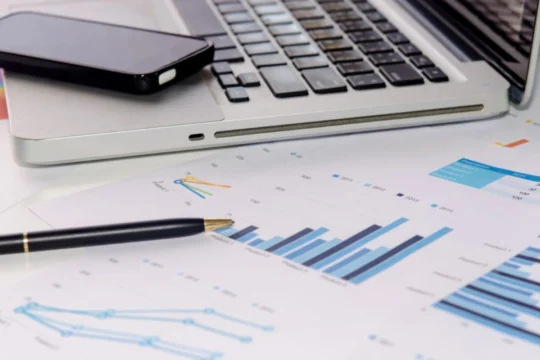 Celular, laptop, papéis e caneta sobre uma mesa, representando produtos financeiros.