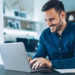 Imagem de um homem sorridente usando notebook, para ilustrar um texto sobre como se tornar correspondente bancário.