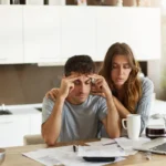 Um casal olha preocupado para as faturas que estão sobre a mesa, ilustrando um artigo da BKOpen sobre como conseguir empréstimo negativado e score baixo.
