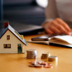 Maquete de uma casa sobre uma mesa, próxima a algumas moedas, representando um artigo da BKOpen sobre como funciona um financiamento.