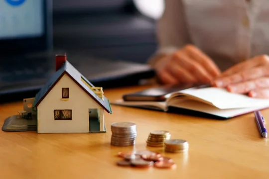 Maquete de uma casa sobre uma mesa, próxima a algumas moedas, representando um artigo da BKOpen sobre como funciona um financiamento.