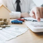 Imagem de uma mão masculina clicando em uma calculadora com vários papéis, moedas e miniaturas de carros e casa ao redor, ilustrando um artigo da BKOpen sobre o que é consórcio.