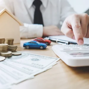 Imagem de uma mão masculina clicando em uma calculadora com vários papéis, moedas e miniaturas de carros e casa ao redor, ilustrando um artigo da BKOpen sobre o que é consórcio.