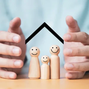 Mão envolvem três bonecos de madeira, que estão apoiados sobre uma mesa, com um telhado desenhado mais acima, representando um artigo da BKOpen sobre seguro de vida como funciona.