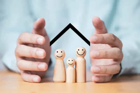 Mão envolvem três bonecos de madeira, que estão apoiados sobre uma mesa, com um telhado desenhado mais acima, representando um artigo da BKOpen sobre seguro de vida como funciona.