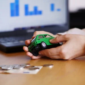 Mãos segurando um carrinho verde com moedas e cédulas ao lado e um laptop com gráficos ao fundo, representando um artigo da BKOpen sobre auto equity.