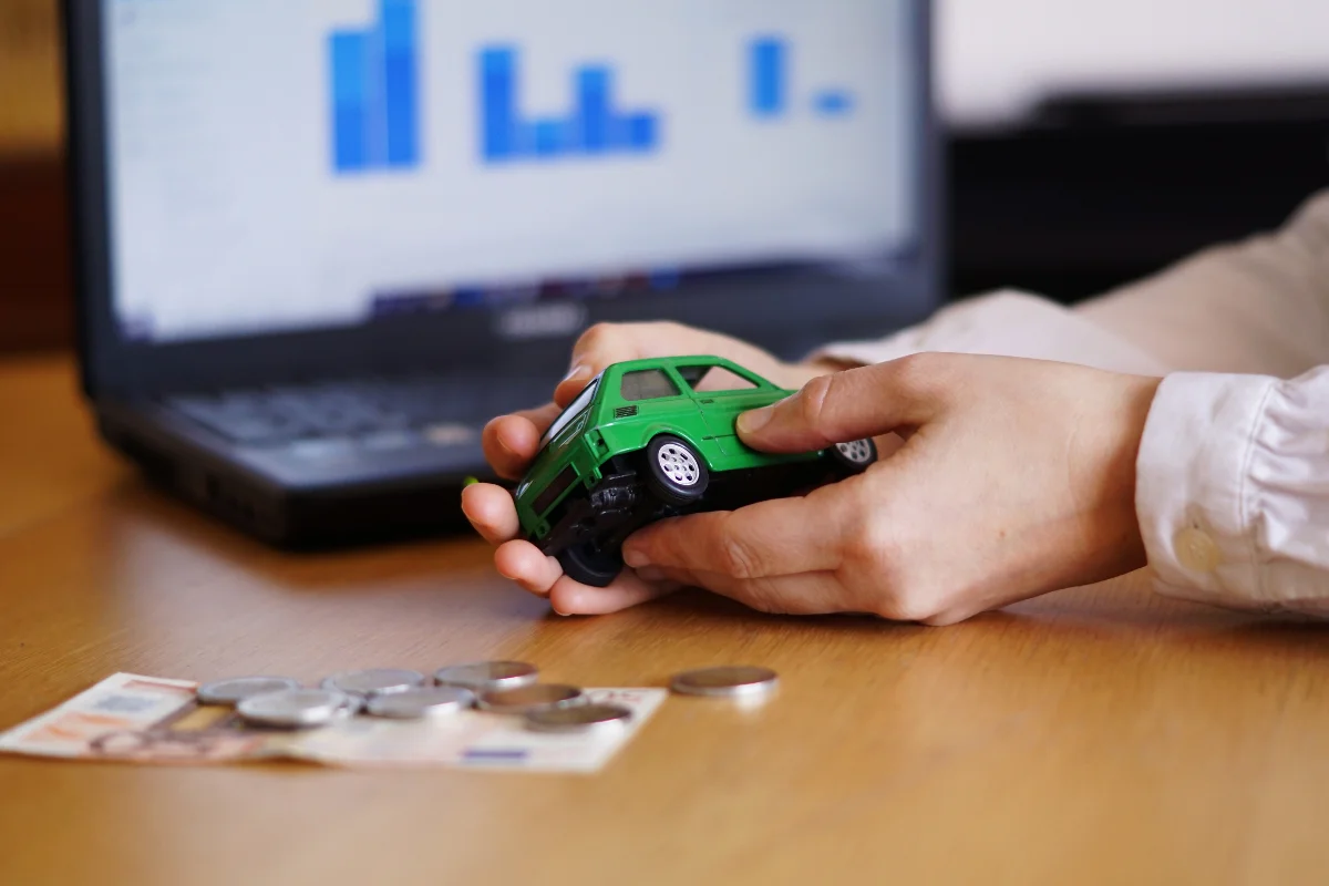 Mãos segurando um carrinho verde com moedas e cédulas ao lado e um laptop com gráficos ao fundo, representando um artigo da BKOpen sobre auto equity.
