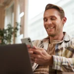Homem sorridente usando smartphone e laptop em ambiente interno com luz natural, ilustrando um artigo da BKOpen sobre empréstimo consignado.