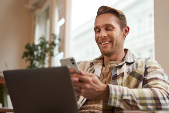 Homem sorridente usando smartphone e laptop em ambiente interno com luz natural, ilustrando um artigo da BKOpen sobre empréstimo consignado.