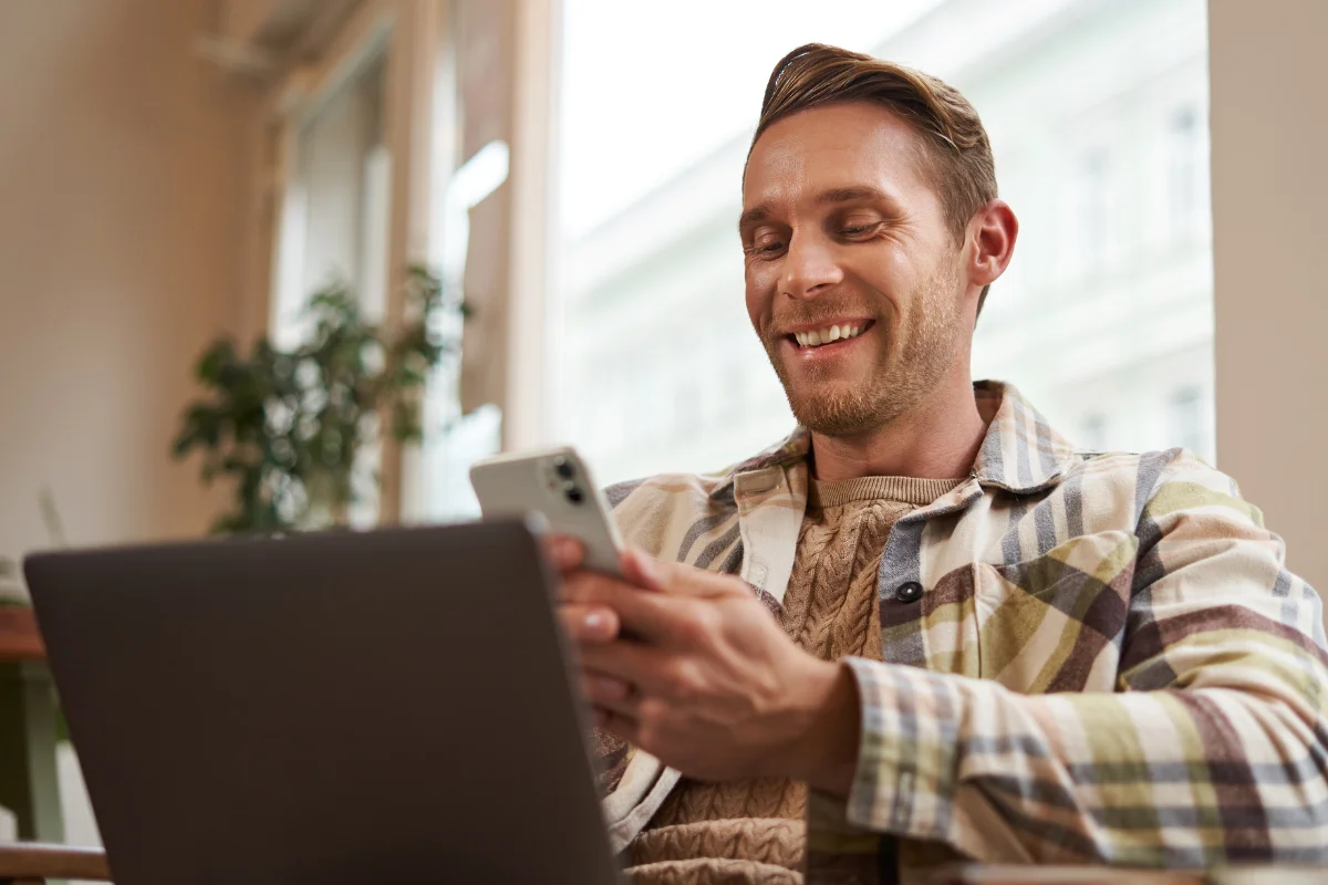 Homem sorridente usando smartphone e laptop em ambiente interno com luz natural, ilustrando um artigo da BKOpen sobre empréstimo consignado.