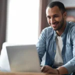 Homem sorridente em casa usando laptop, com xícara de café na mesa, ilustrando um artigo da BKOpen sobre marketplace financeiro.