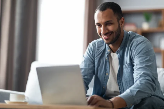 Homem sorridente em casa usando laptop, com xícara de café na mesa, ilustrando um artigo da BKOpen sobre marketplace financeiro.