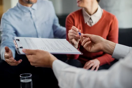Duas pessoas assinam um contrato com um consultor financeiro, ilustrando um artigo da BKOpen sobre o que é preciso para fazer um consórcio.