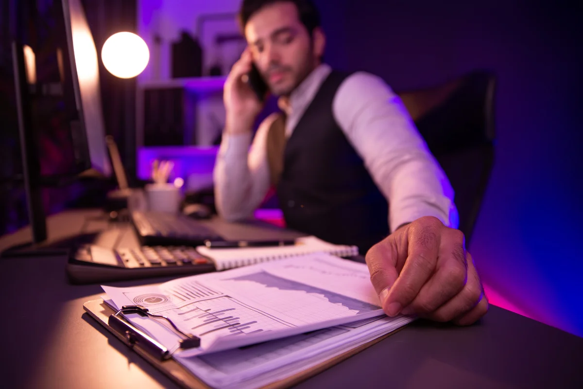 Imagem de homem falando ao telefone enquanto analisa gráficos financeiros em escritório com iluminação roxa, ilustrando um artigo da BKOpen sobre refinanciamento de empréstimo consignado.