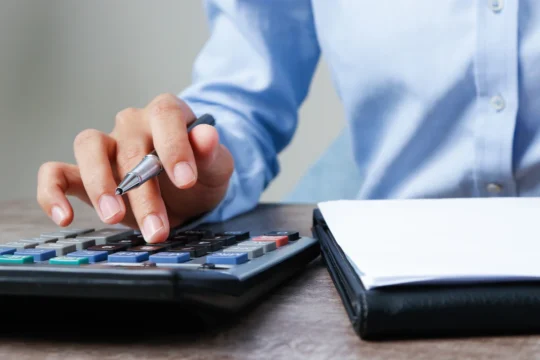 Pessoa usando calculadora com caneta na mão e caderno ao lado em mesa de madeira, ilustrando um artigo da BKOpen sobre como calcular juros de financiamento.