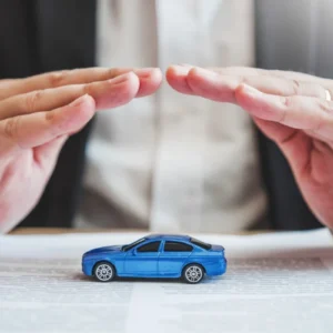 Mãos protegendo um carro de brinquedo azul sobre um documento, simbolizando como funciona um seguro de carro em artigo da BKOpen.