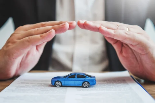 Mãos protegendo um carro de brinquedo azul sobre um documento, simbolizando como funciona um seguro de carro em artigo da BKOpen.