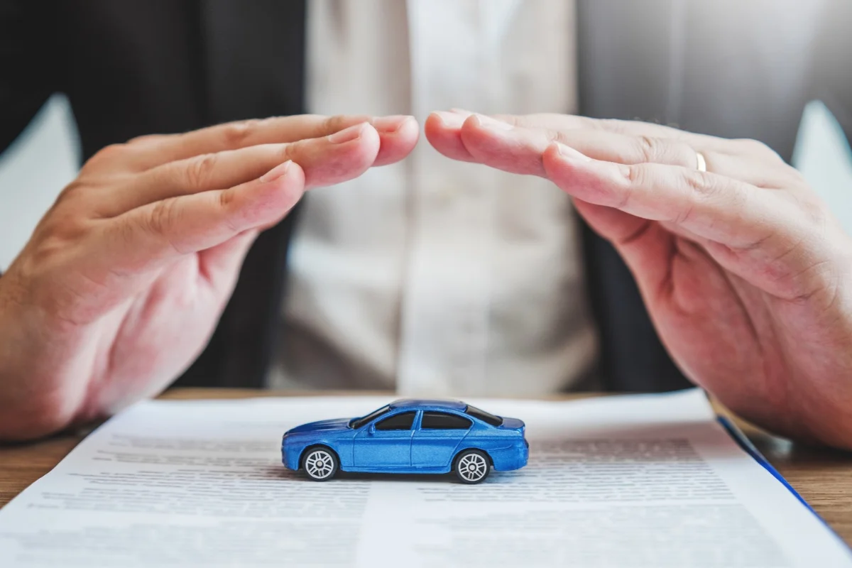 Mãos protegendo um carro de brinquedo azul sobre um documento, simbolizando como funciona um seguro de carro em artigo da BKOpen.