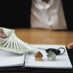 Pessoa segurando dinheiro em frente a miniaturas de casas e chave sobre caderno aberto, ilustrando um artigo da BKOpen sobre qual a diferença entre consórcio e financiamento.