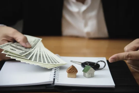 Pessoa segurando dinheiro em frente a miniaturas de casas e chave sobre caderno aberto, ilustrando um artigo da BKOpen sobre qual a diferença entre consórcio e financiamento.