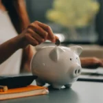 Pessoa colocando moeda em cofrinho branco ao lado de calculadora e caderno laranja em mesa, ilustrando um conteúdo da BKOpen sobre o que é educação financeira.