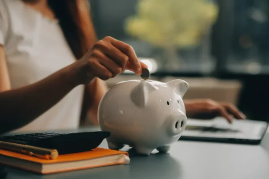 Pessoa colocando moeda em cofrinho branco ao lado de calculadora e caderno laranja em mesa, ilustrando um conteúdo da BKOpen sobre o que é educação financeira.